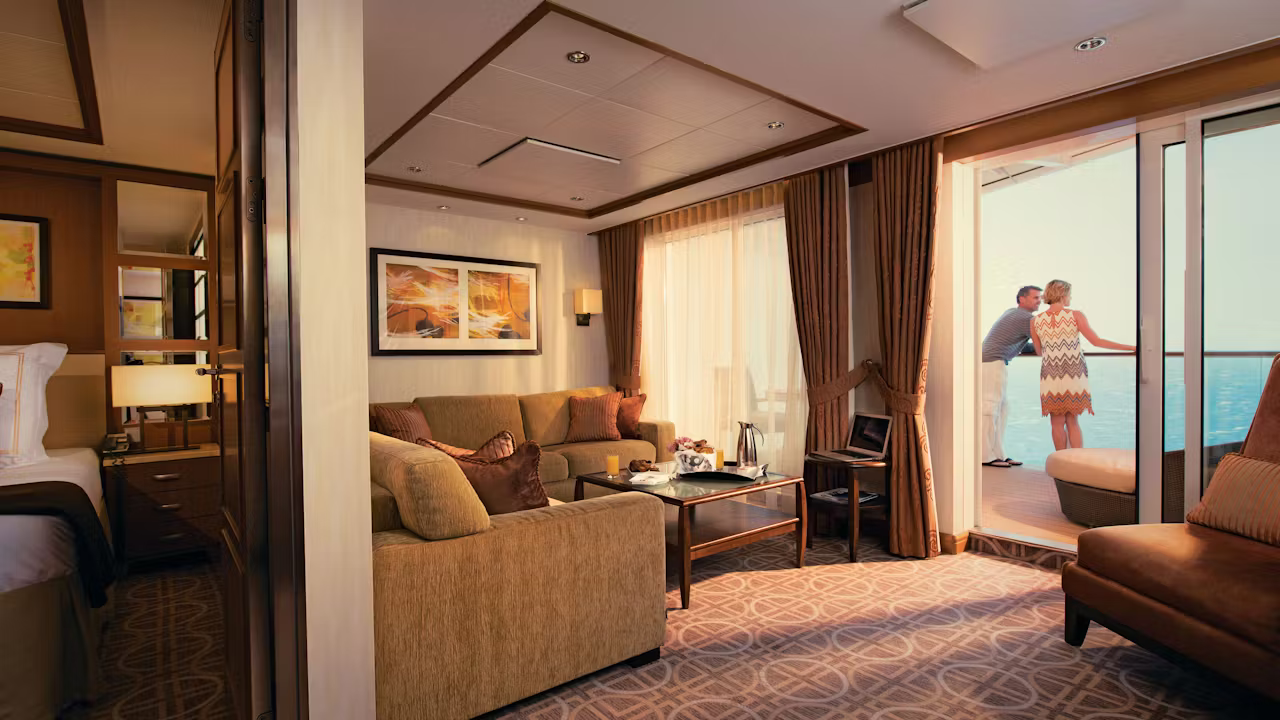 Ruime cruisesuite met aparte zithoek, slaapkamer en balkon met uitzicht op zee waar twee gasten ontspannen aan de reling staan. 