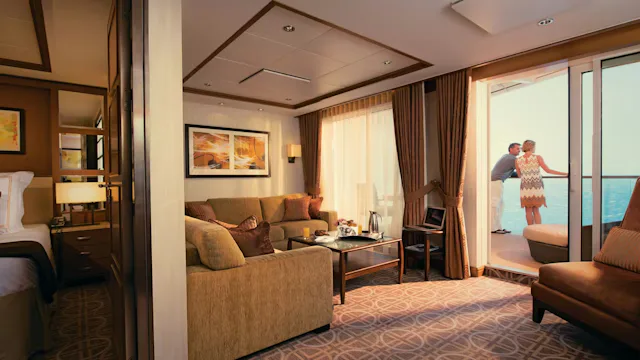 Ruime cruisesuite met aparte zithoek, slaapkamer en balkon met uitzicht op zee waar twee gasten ontspannen aan de reling staan.