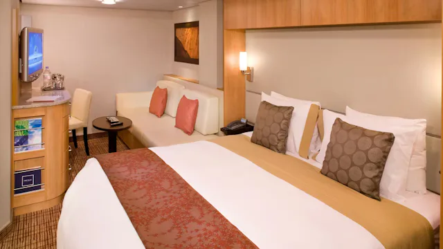 Ruime cruisehut met tweepersoonsbed, zithoek met bank en tafel, bureau met televisie en warme houten accenten in het interieur.