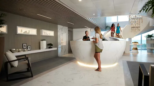 Gast die wordt geholpen bij een moderne receptiebalie in een stijlvolle lounge- of serviceomgeving aan boord van een cruiseschip.
