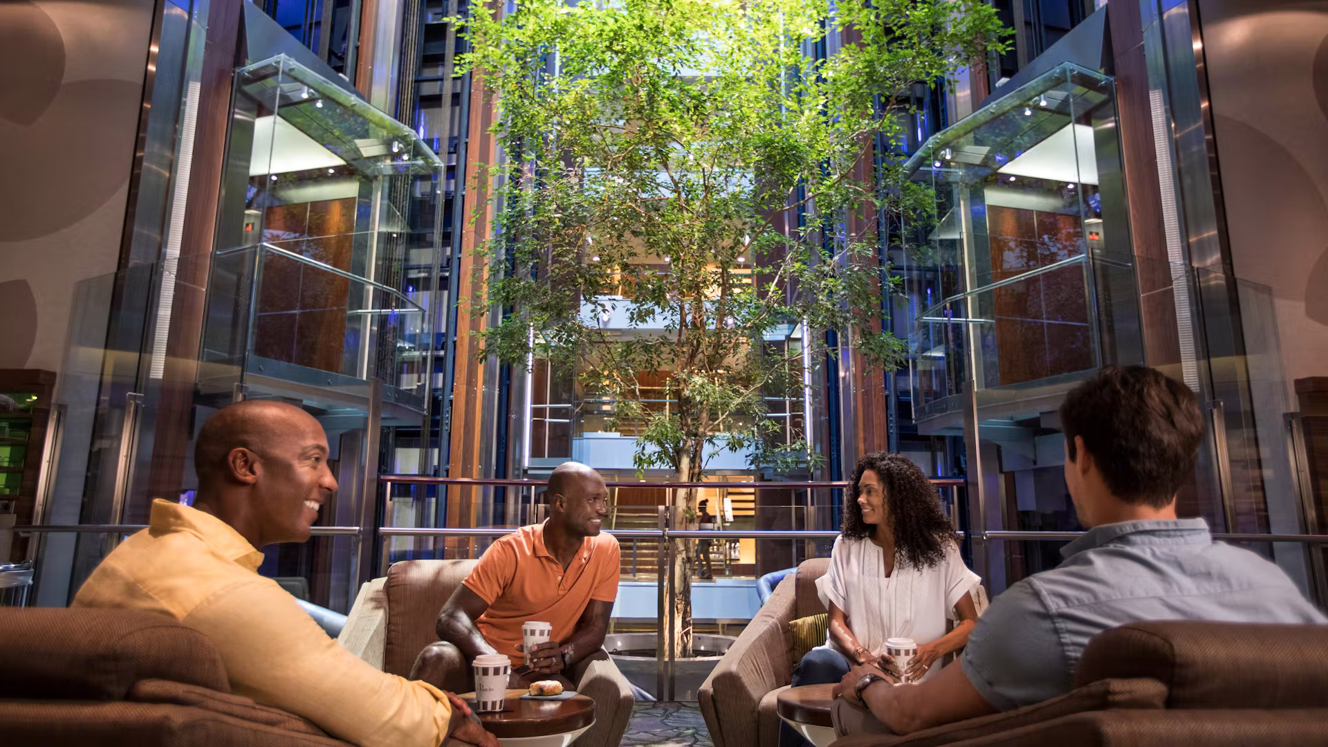 Gezellige lounge aan boord van een cruiseschip waar vier gasten ontspannen samen zitten met koffie, omringd door een modern interieur met glazen liften en een grote boom