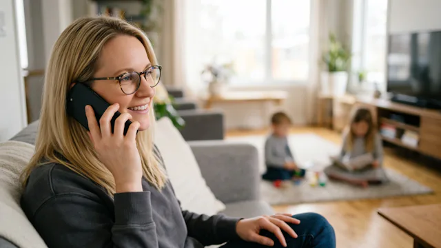 Vrouw belt glimlachend vanuit haar woonkamer met de klantenservice over haar cruiseboeking, terwijl haar kinderen op de achtergrond spelen.